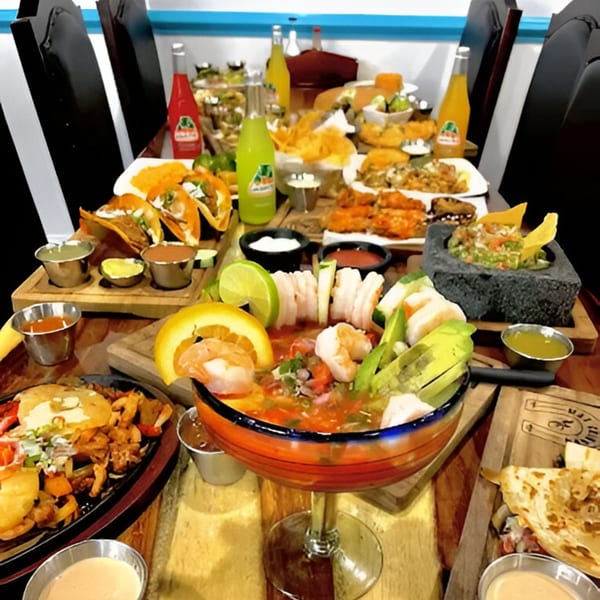A table laden with various Mexican dishes, including tacos, ceviche, guacamole, and Jarritos sodas.