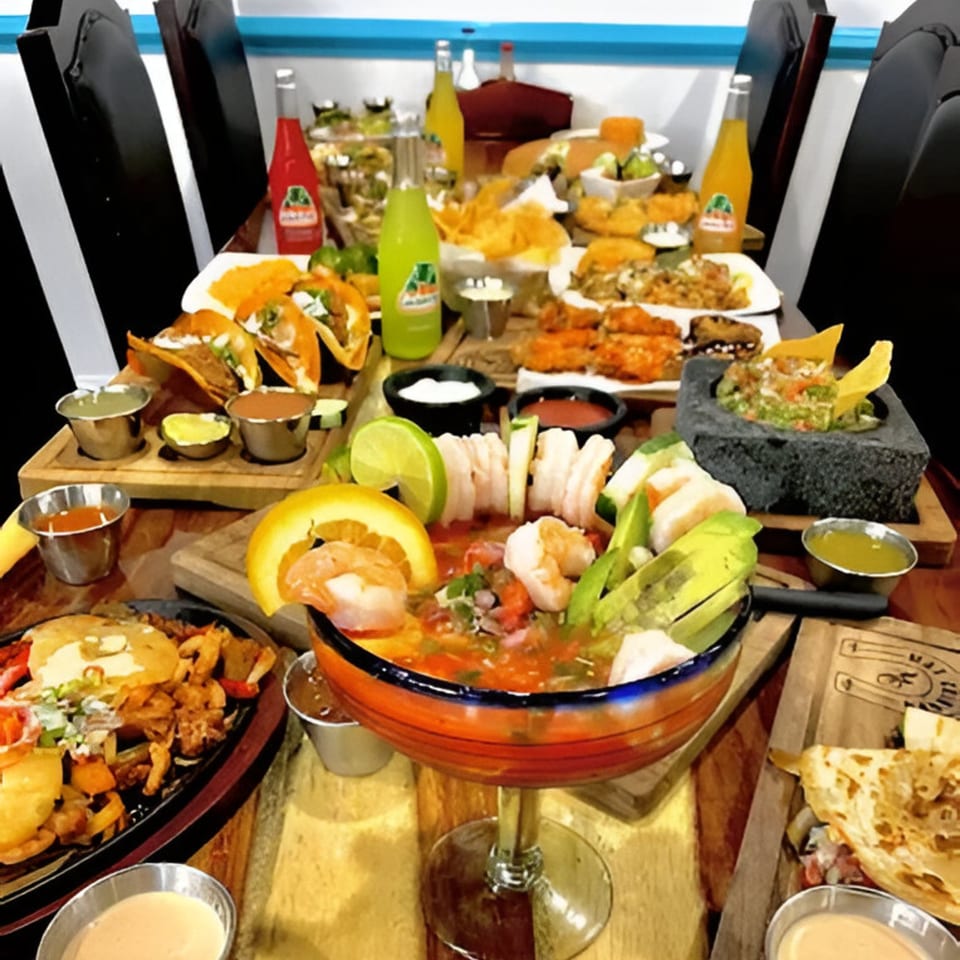 A table laden with various Mexican dishes, including tacos, ceviche, guacamole, and Jarritos sodas.