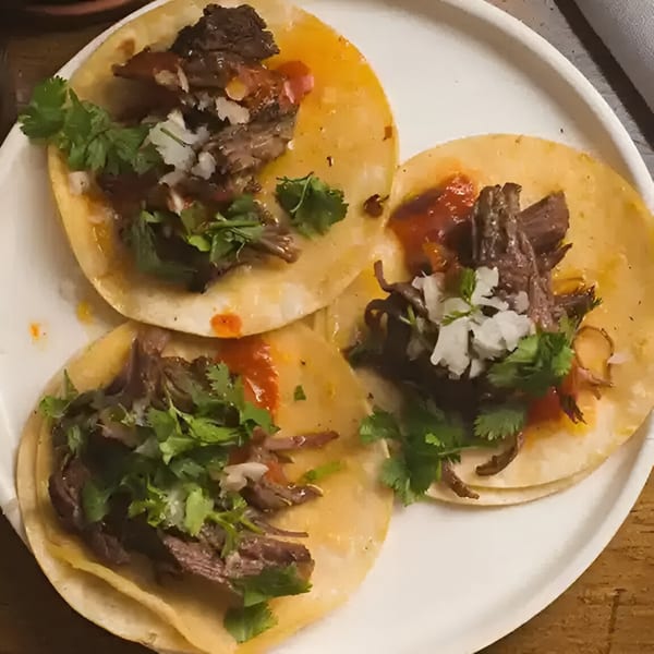 Three Mexican tacos with shredded meat, red sauce, diced onions, and cilantro on a white plate.