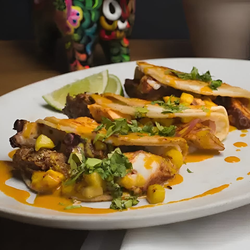 Three tacos with mango salsa, cilantro, and orange sauce, served with lime wedges on a white plate.