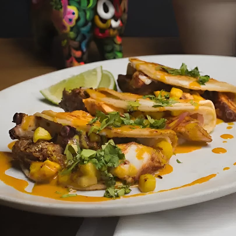 Three tacos with mango salsa, cilantro, and orange sauce, served with lime wedges on a white plate.