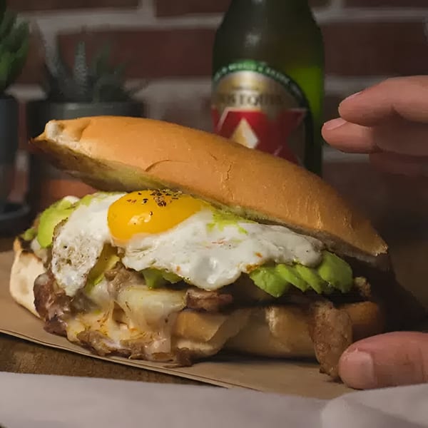 A Mexican torta sandwich with steak, avocado, and a fried egg, with a beer bottle in the background.