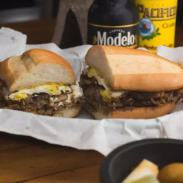 Two Mexican torta sandwiches with meat and eggs, with Modelo and Pacifico beer bottles in the background.