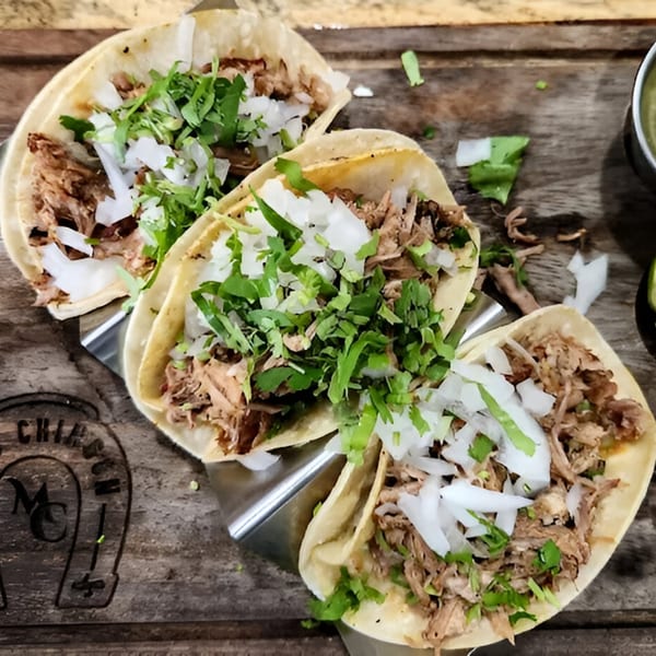 Three carnitas tacos with onions and cilantro on a wooden board with a branded horseshoe.